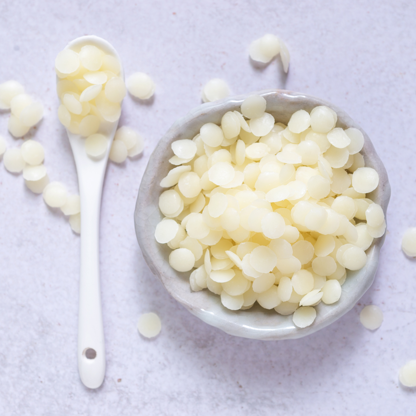 Waxy white beads of btms-225 in a bowl with a spoon on a light gray background