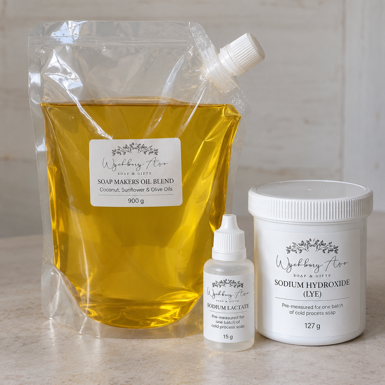 Three products from Wychbury Ave Soap & Gifts arranged on a light stone surface: a large clear pouch of soap maker's oil blend, a small bottle of potassium lactate, and a white tub of sodium hydroxide (lye).