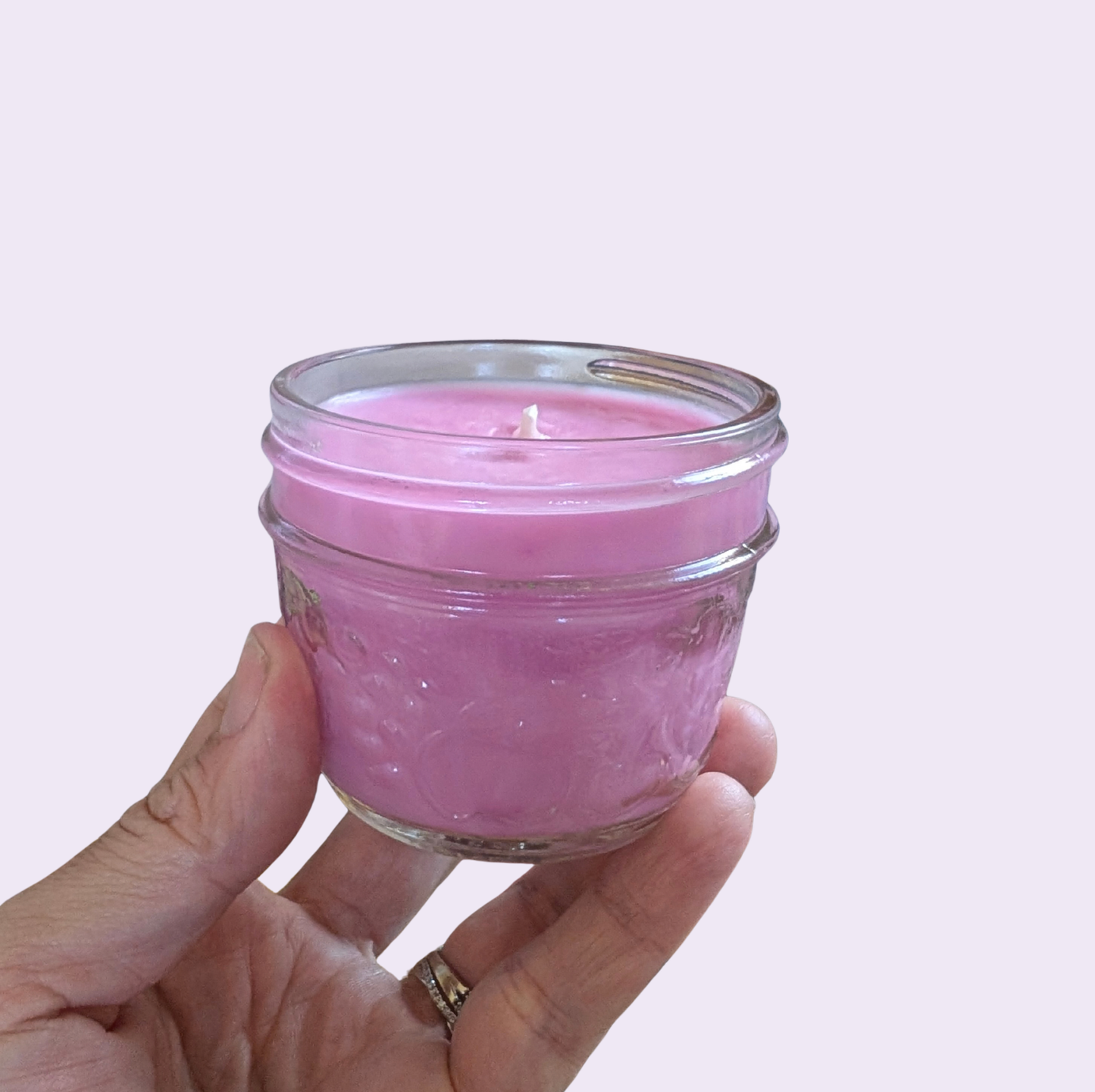 A hand holding a small pink candle in a clear glass jar against a solid pale pink background.