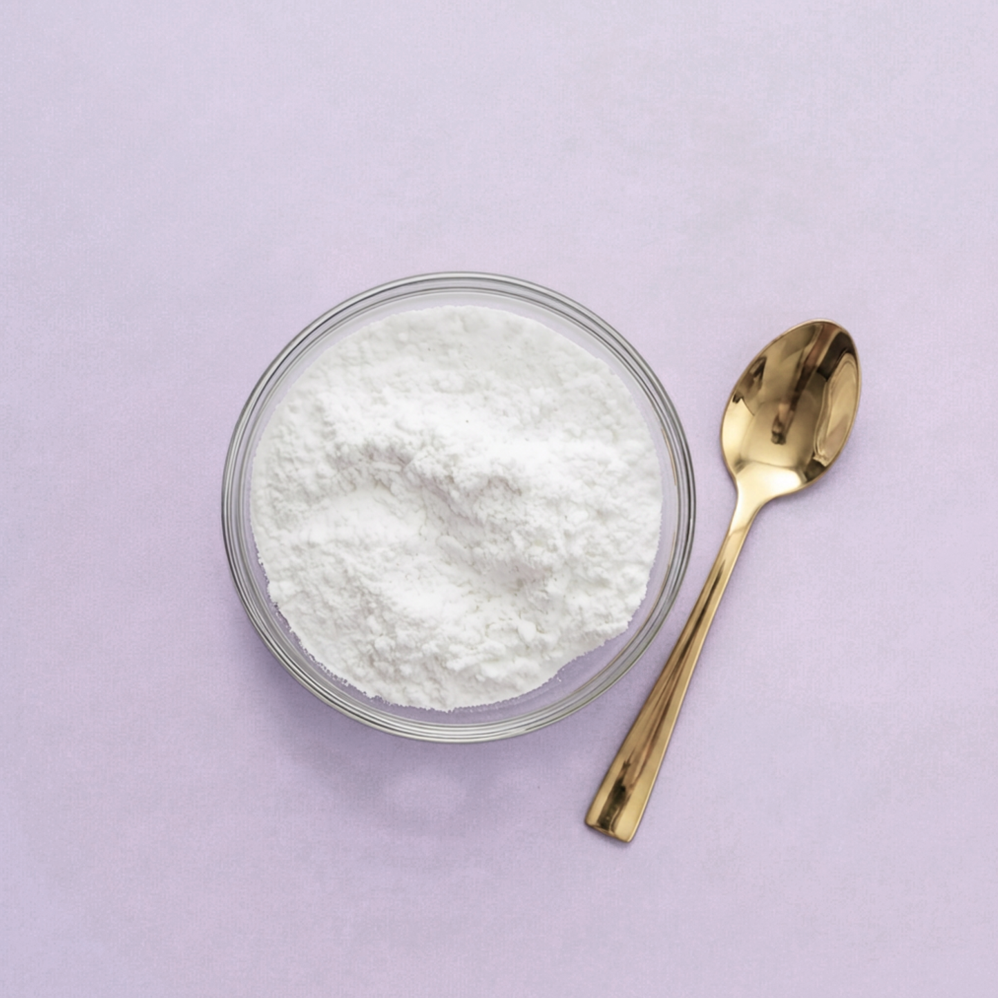 Clear glass bowl with white powder and a gold spoon on a light purple background