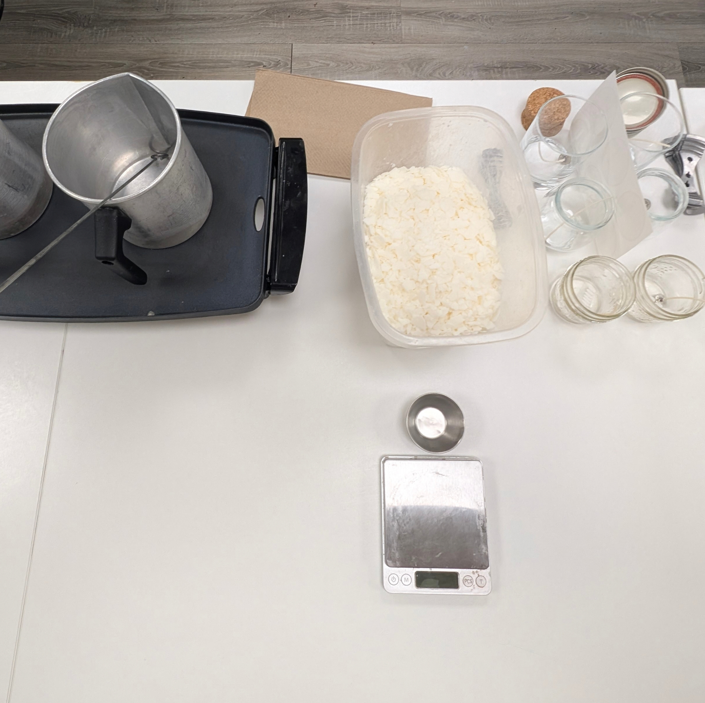A top-down view of a workspace on a white table showing a digital scale, a metal cup on a black tray, a container of white soy wax flakes, glass jars, and a small electronic device.