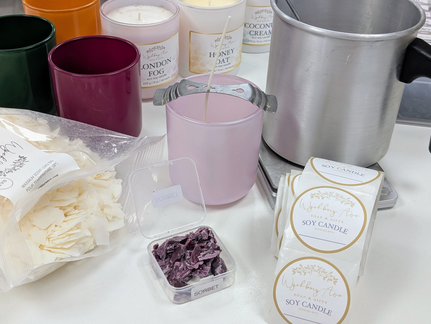 Soy candle making supplies and finished candles arranged on a white surface, including wax melts, a pink container with a wick, labeled soy candle packs, colorful cups, and a metal pot.