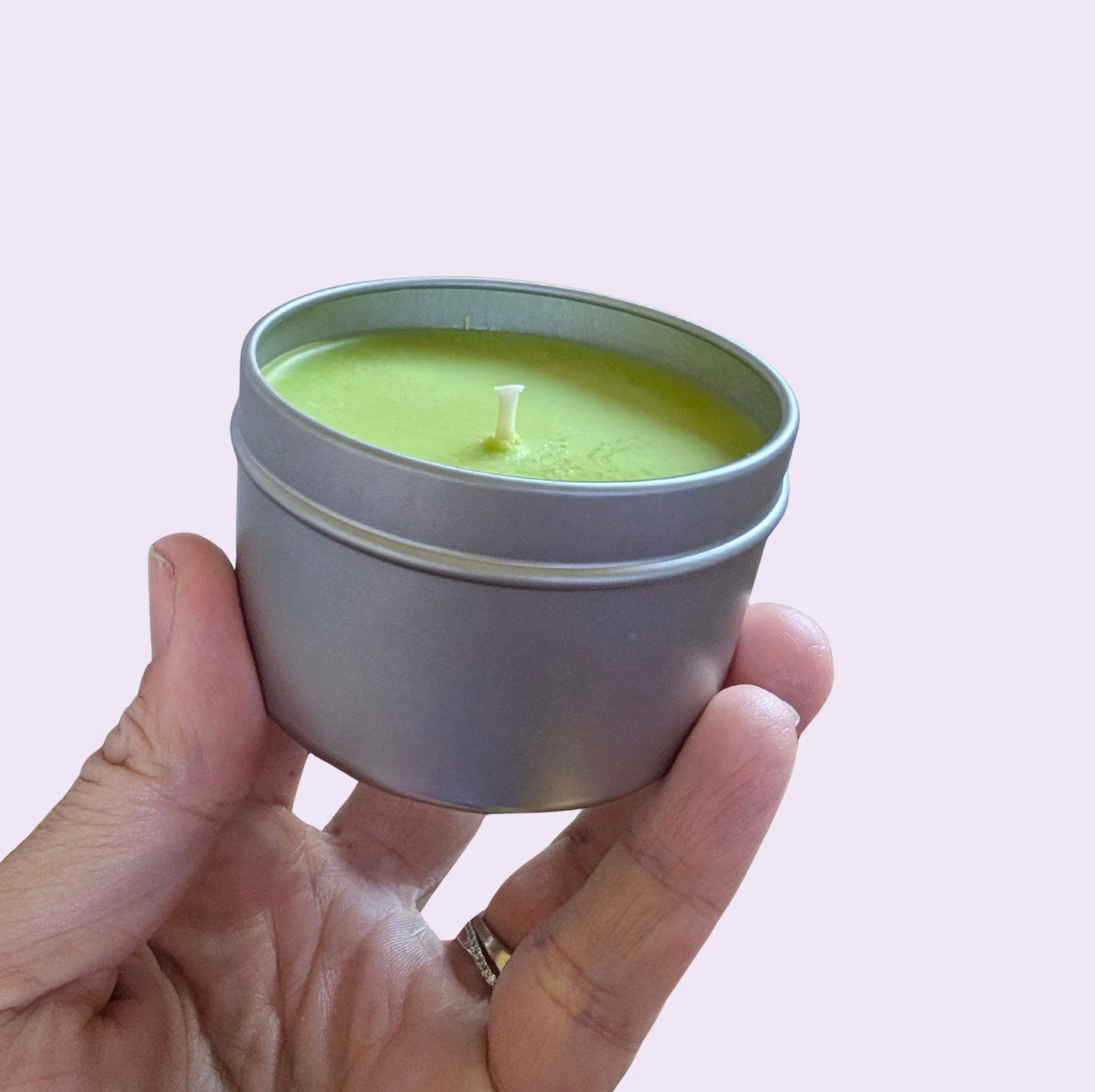 A hand holding a small round silver metal tin candle with light green wax and a single white wick, set against a solid pale pink background.