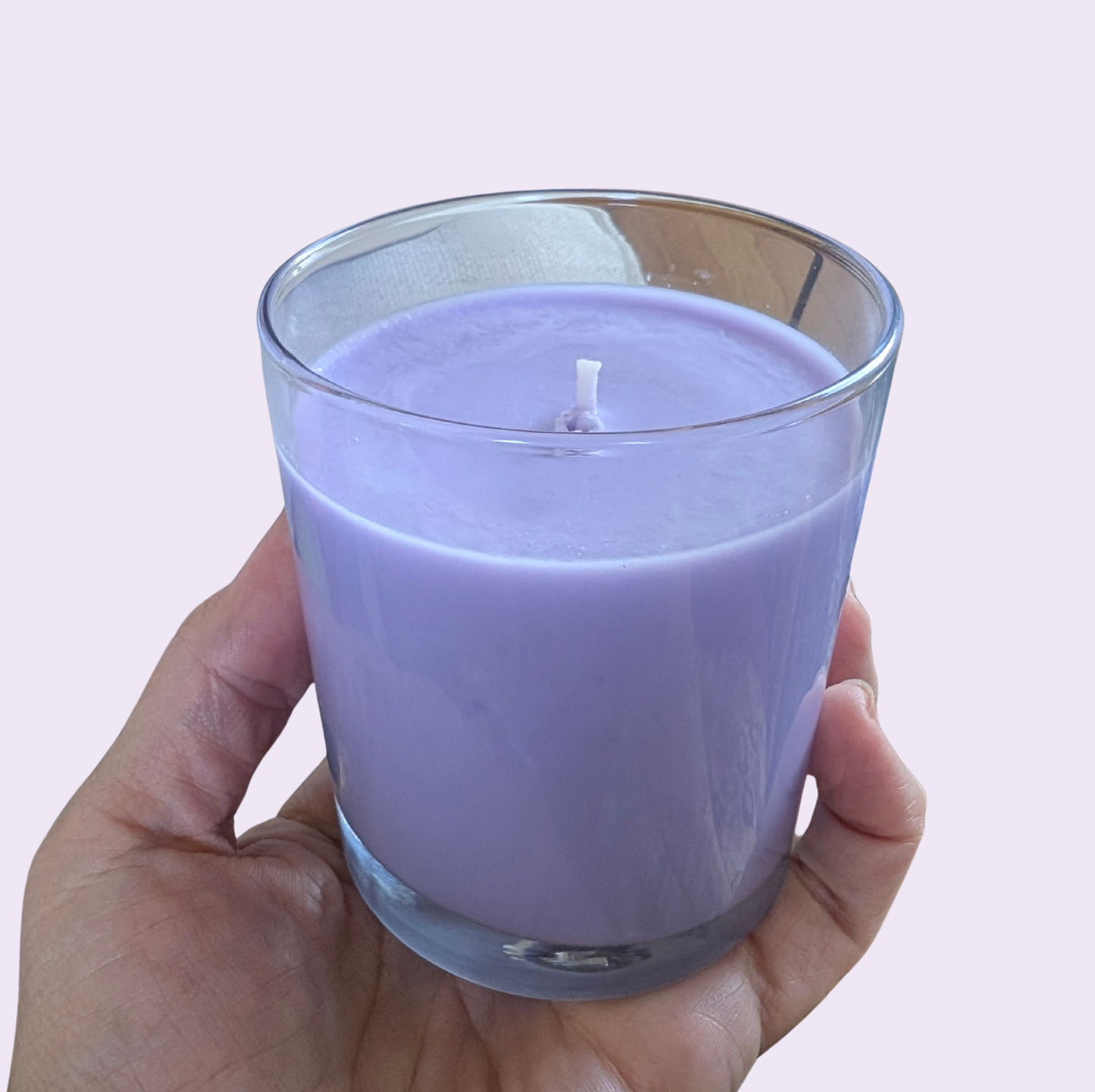 A hand holding a lavender purple candle in a clear glass jar against a solid pale pink background.